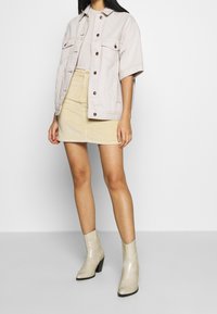 Giacca di jeans beige chiaro oversize con due tasche sul petto, abbinata a una minigonna in velluto a coste giallo texturizzato e stivaletti crema lucidi.