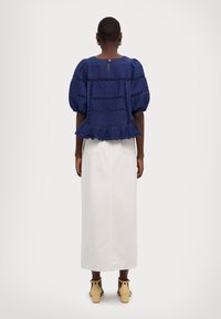 Top blu navy con maniche a sbuffo in tessuto strutturato, orlo arricciato e apertura a goccia sulla schiena, abbinato a una gonna lunga color avorio e sandali gialli con tacco a blocco.