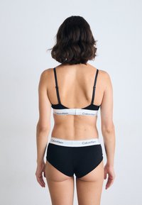 Donna con capelli scuri di lunghezza spalle, che indossa lingerie bianca e nera di Calvin Klein, in piedi di spalle contro uno sfondo chiaro e neutro.