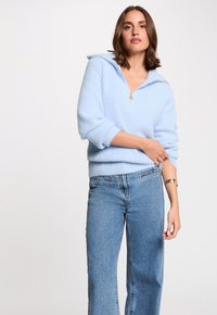 Suéter azul claro de gran tamaño en tejido suave y peludo con cuello con cremallera; combinado con jeans azul de cintura alta. Textura suave, ajuste relajado.