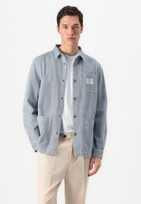 Jaqueta camisa de denim riscada em azul e branco, com botão à frente, bolso no peito e dois bolsos inferiores. Textura suave, ajuste casual.
