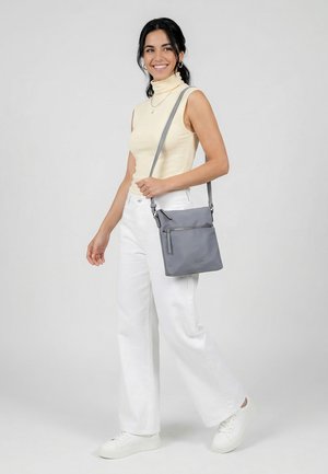 Smiling woman wearing sleeveless cream turtleneck and white pants, carrying small gray crossbody bag and white sneakers, standing on gray background.