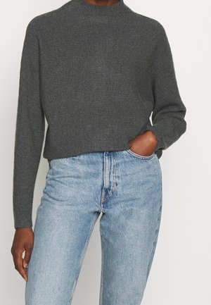 Persoon draagt een donkergrijze geribbelde trui met lange mouwen en lichtblauwe high-waisted jeans, met de rechterhand in de zak, tegen een witte achtergrond.