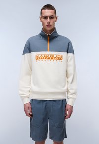 Light cream and gray pullover with a zip collar, featuring orange branding. Worn with navy blue, textured shorts with large pockets.