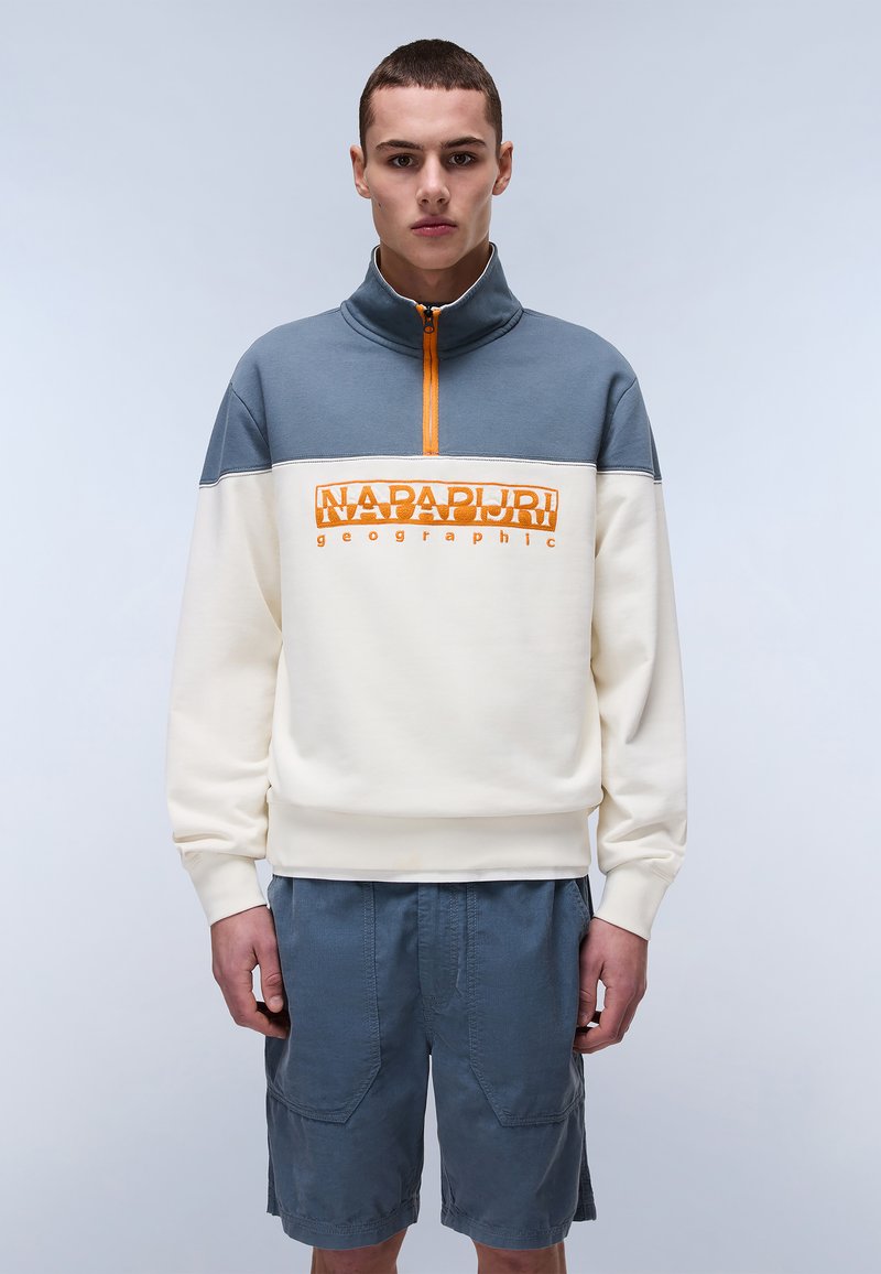 Light cream and gray pullover with a zip collar, featuring orange branding. Worn with navy blue, textured shorts with large pockets.