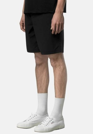 Uomo che indossa pantaloncini neri, calzini bianchi e scarpe da ginnastica in tela bianche, in piedi davanti a uno sfondo semplice.