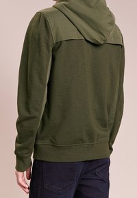 Olivengrøn hættetrøje lavet af blødt stof med tekstureret øverste panel, justerbar hætte og ribbet kant. Bæres med mørke jeans.