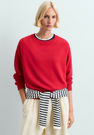 Lyshåret person iført en rød langærmet top med en sort-hvidstribet sweater bundet om livet og lysfarvede bukser.
