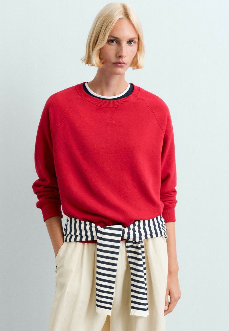Personne blonde portant un haut rouge à manches longues avec un pull rayé noir et blanc noué autour de la taille et un pantalon de couleur claire.