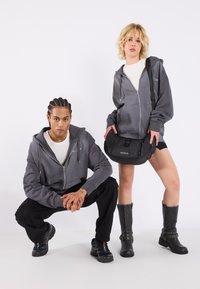Siva zip-up kapuca, bela majica, črne hlače, športni čevlji in črna torba za ramo z sponko; model pozira knežeč, drugi pa stoji.