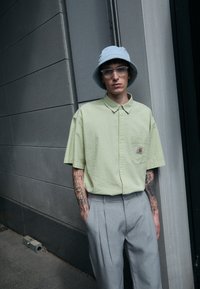 Korte mouwen, lichtgroen, gestructureerd overhemd met knopen en een borstzak; gecombineerd met lichtgrijze, geplooide broek en een lichtblauwe bucket hat.