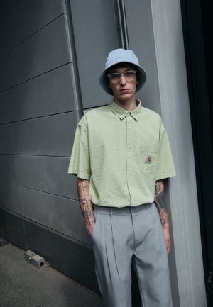 Chemise à manches courtes, vert clair, texturée avec un poche poitrine ; associée à un pantalon léger gris clair plissé et un chapeau seau bleu clair.