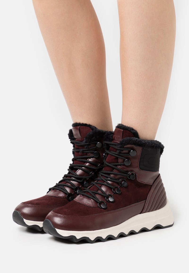 Stivali alla caviglia color marrone scuro con fodera in pelliccia nera, suede texturizzato e pelle liscia. Presentano lacci neri e una suola bianca zigzag.