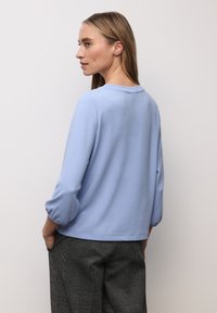 Jasnoniebieska bluzka z długim rękawem, okrągłym dekoltem i bufkami na rękawach, w zestawieniu z czarno-białymi wzorzystymi spodniami. Gładka struktura tkaniny.