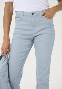 Pantalon en denim bleu clair avec une coupe ajustée, couture visible et poches avant, porté avec un haut blanc. Main tenant une veste.