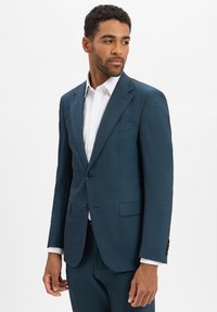 Donkergroen pak met een gestructureerde stof, enkelbreasted ontwerp, met gesneden revers en twee knopen aan de voorkant. Witte shirt eronder.