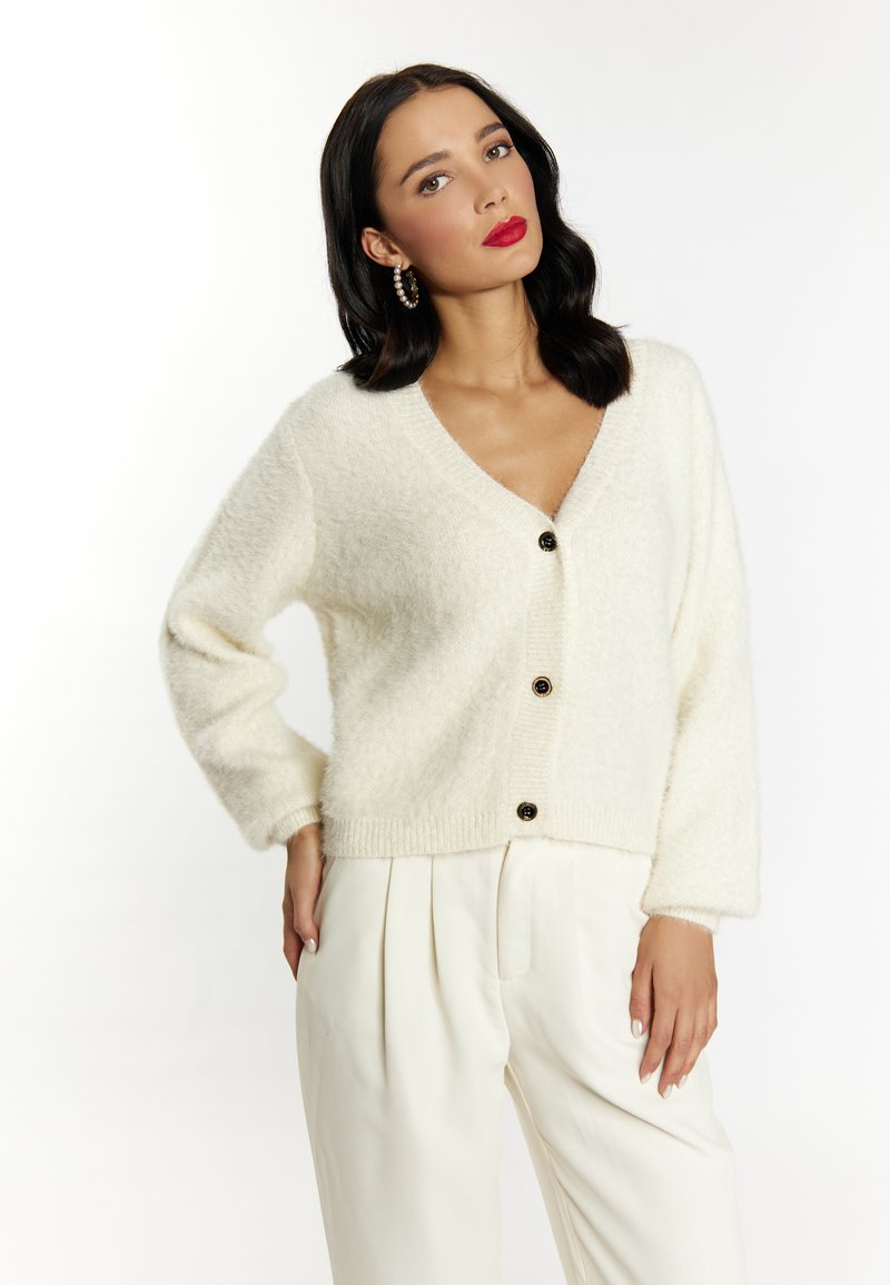 Cardigan fuzzy color crema con scollatura a V, maniche lunghe e quattro bottoni neri. Indossato sopra pantaloni bianchi a pieghe.