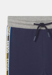 Námořnické joggers s šedým žebrovaným pasem a šňůrkou. Vybaven bílými a barevnými pruhy po stranách s brandingem "GUESS". Měkký materiál.