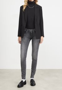Blazer negro sobre un cuello de tortuga negro, combinado con jeans gris desgastados ajustados y mocasines negros. Un collar de cadena simple acentúa el escote.