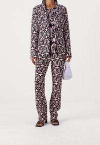 Costume à motifs en noir et rose avec un design floral. La veste est ajustée, accompagnée de pantalons assortis. Accessoirisé avec un petit sac blanc.