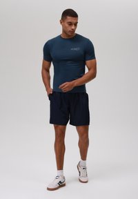 Camiseta ajustada de color azul marino en material suave con mangas cortas, combinada con pantalones cortos oscuros. El calzado presenta detalles en blanco y azul marino.