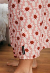 Rožinė audinio medžiaga su geometrinėmis formomis, papuošta raudonais akcentais ir baltais deimantais. Etiketė matoma šalia krašto, ant medinių grindų fono.