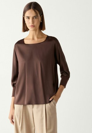 Frau mit glatten, schulterlangen braunen Haaren, die eine dunkelbraune lockere Bluse mit Dreiviertelärmeln und beige Hosen vor einem einfarbigen Hintergrund trägt.