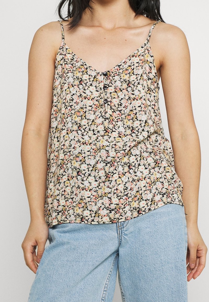 Femme portant un haut sans manches à motif floral avec des boutons, assorti à un jean denim bleu clair, sur un fond uni.