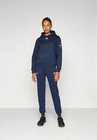 Sweatshirt azul-marinho com capuz, bolso canguru e cordão, combinado com calças de fato de treino correspondentes, apresentando logótipos, tecido texturizado e punhos ajustados.
