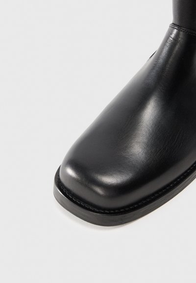 Bota de cuero negro hasta el tobillo con punta redondeada, textura suave y una suela de goma negra resistente. Detalle de costura mínima a lo largo del borde.