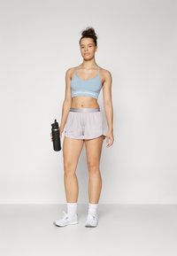 Lichtblauwe sportbeha met een logo, grijze shorts met een elastische tailleband en een zwarte waterfles. Model draagt witte sportschoenen en sokken.