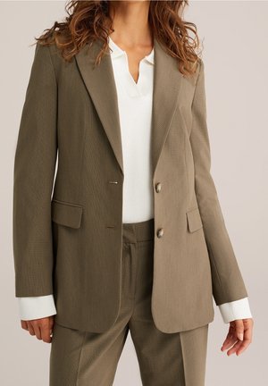 Femme portant un blazer marron bicolore sur mesure avec un pantalon assorti et un pull blanc côtelé en dessous.