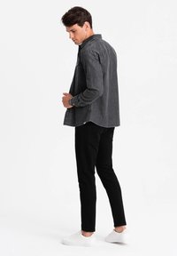 Zwart denim overhemd met lange mouwen, knoopmanchetten en een gebogen zoom, gedragen over zwarte broek en witte sneakers.