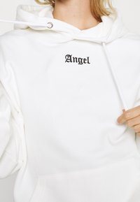 Bílá mikina s kapucí vyrobená z měkké tkaniny, s přední kapsou, nastavitelnými šňůrkami a slovem "Angel" vyšitým černým kurzivním písmem.