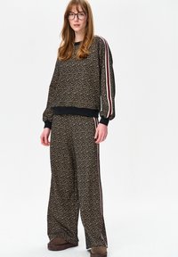 Leopardprint trainingspak van zachte stof met elastische manchetten, gestreepte zijstrepen in bordeauxrood en wit, en broek met wijde pijpen. Bruine schoenen.