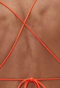 Oranžové prekrížené ramienka na opálenej pokožke, na chrbte sťahované mašľou. Hladká textúra, tenké ramienka s minimalistickým dizajnom.