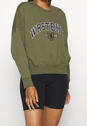 Osoba w oliwkowej bluzie z napisem „WRSTBHN” i herbem, w połączeniu z czarnymi dopasowanymi szortami kolarskimi.