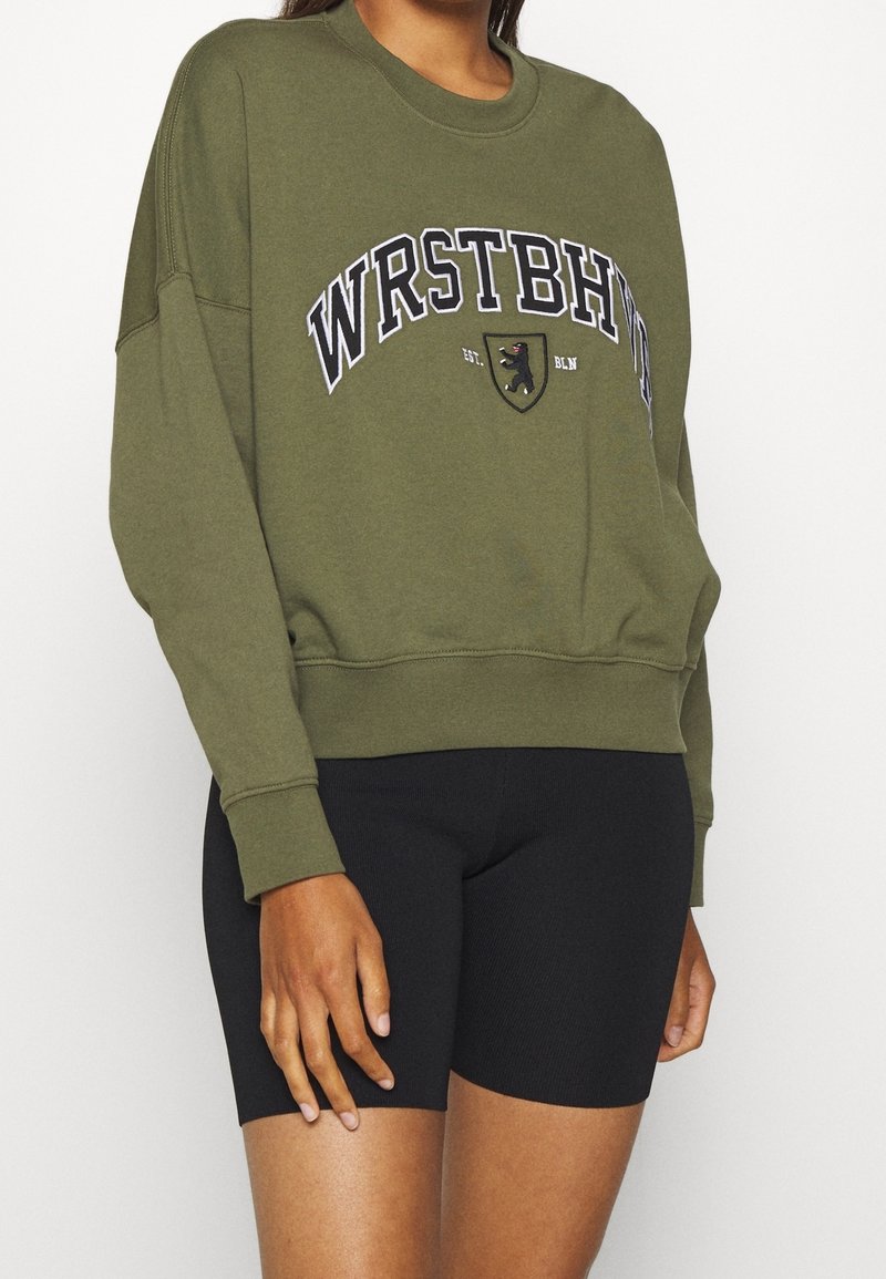 Personne portant un sweat-shirt vert olive avec le texte « WRSTBHN » et un blason, associé à un short de cyclisme noir ajusté.