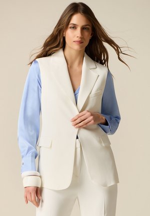 Frau mit langen braunen Haaren, die ein hellblaues Hemd unter einem ärmellosen weißen Blazer und passenden weißen Hosen trägt, steht vor einem beigen Hintergrund.