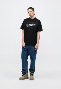 Czarna bawełniana koszulka z metalicznym, srebrnym logo "Carhartt", zestawiona z luźnymi niebieskimi dżinsami i oliwkowo-zielonymi sznurowanymi butami.