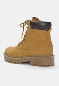 Stivale alla caviglia in pelle tan con punta arrotondata, distintivi dettagli in similpelle marrone e una suola in gomma spessa e texturizzata. Presenta occhielli in metallo e lacci.