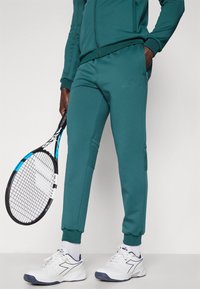 Teal joggebukser laget av mykt stoff, med glidelåslommer og ribbestrikkede mansjetter. Kombinert med hvite joggesko og holder en tennisracket.