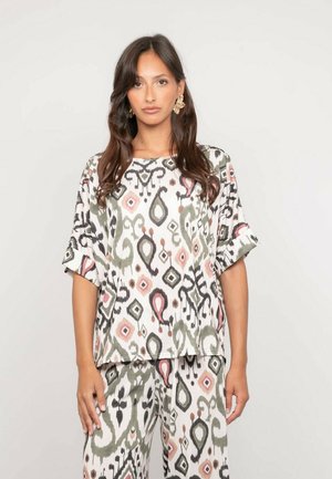 Femme aux longs cheveux bruns portant un haut ample et un pantalon assortis avec un imprimé abstrait noir, vert, rose et beige sur un fond clair.