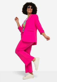 Blazer rose et pantalon assorti en tissu lisse, associé à un t-shirt graphique. Le mannequin porte des baskets blanches et des lunettes de soleil rondes.