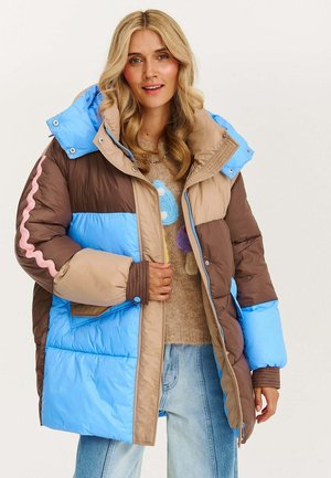 Kobieta z długimi blond włosami w kolorowej kurtce puchowej w niebieskim, brązowym i beżowym kolorze, z wzorzystym swetrem i jasnymi dżinsami.