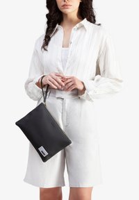 Sort rektangulær clutch-taske med en tekstureret overflade og guld lynlås, holdt af en person iført en hvid stribet bluse og lyse shorts.