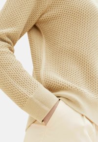 Maglione beige con texture e motivo intrecciato, dotato di polsini a coste e design aderente, indossato sopra pantaloni chiari.