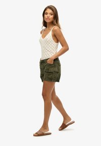 Débardeur en tricot blanc avec un motif en diamant, short cargo vert olive avec plusieurs poches, et sandales plates marron. Modèle en profil latéral.