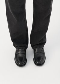 Svarta läderloafers med en slät yta, dekorativ remdetalj och något rundade tår, kombinerade med mörka jeans.