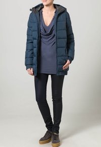 Marineblå pufferjakke med hætte, som har lynlås og elastiske manchetter, kombineret med en draperet grå top og mørke skinny jeans.